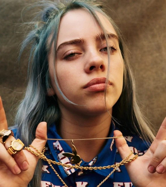 Close-up portrait of a young adult female with dyed blue hair, gold jewelry, and a patterned top against a neutral background.