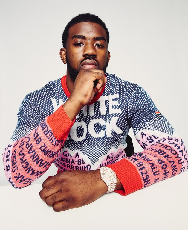 Portrait of a Black male model wearing a patterned White Rock sweater and watch with a neutral expression in a studio setting.