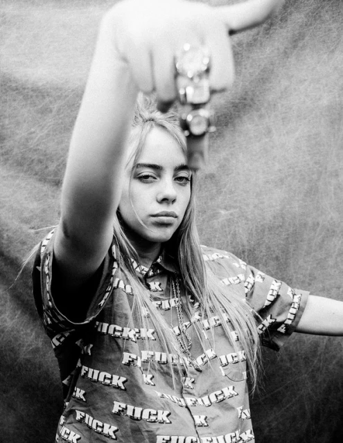 Monochrome portrait of Billie Eilish wearing a shirt with bold 'FUCK' text, hand raised with rings, against a mottled background.
