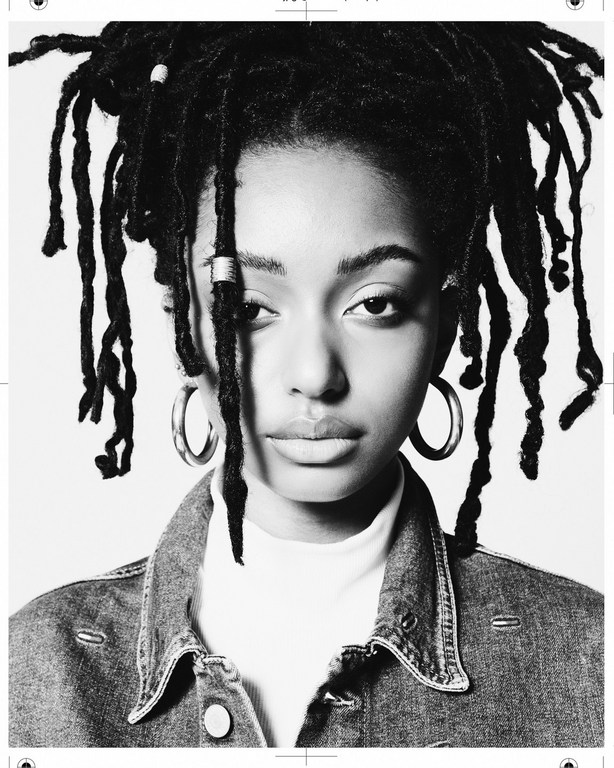 Close-up black-and-white portrait of an adult woman with dreadlocks, hoop earrings, and a denim jacket against a white backdrop.