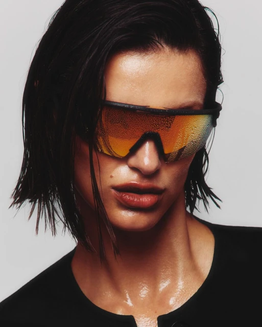 Close-up portrait of a woman with wet dark hair wearing amber-mirrored sport sunglasses against a plain grey studio backdrop.