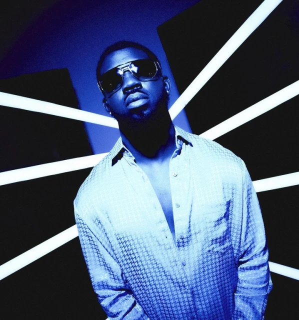 Stylish man in sunglasses and patterned shirt poses against a geometric backdrop in a blue-toned, high-contrast studio setting.