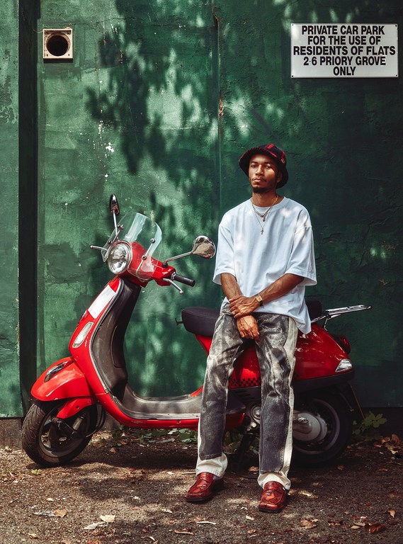 Young man in bucket hat sits on a red scooter in front of a green wall with signage, wearing a white t-shirt and patterned jeans.