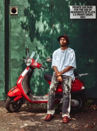 Young man in bucket hat sits on a red scooter in front of a green wall with signage, wearing a white t-shirt and patterned jeans.