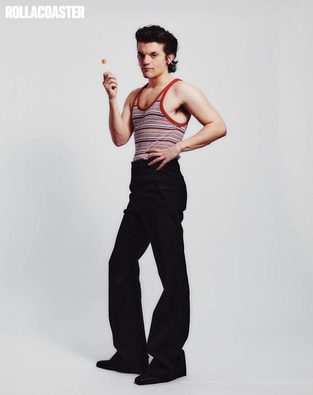 Full-length studio shot of a male model posing with a lollipop. He wears a striped tank top and black trousers against a neutral backdrop.