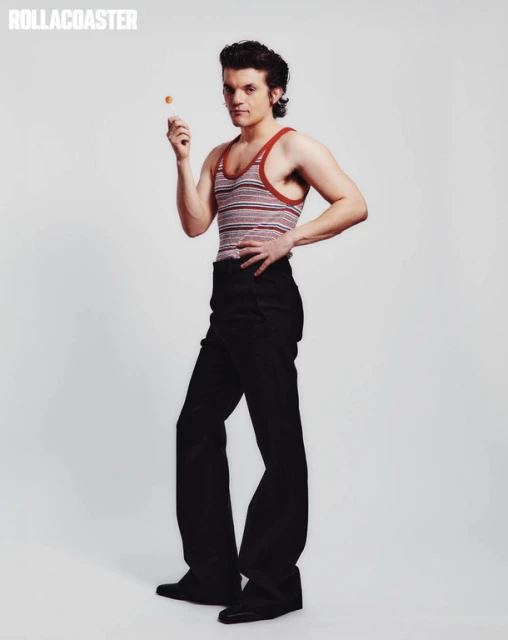 Full-length studio shot of a male model posing with a lollipop. He wears a striped tank top and black trousers against a neutral backdrop.