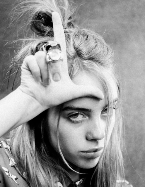 Black and white portrait of a young adult female with blonde hair, wearing a ring and posing with her hand on her forehead.