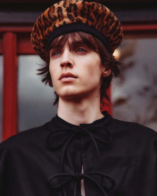 Close-up portrait of a young adult with dark hair, wearing a leopard-print beret and a black top with a bow tie, posing indoors.