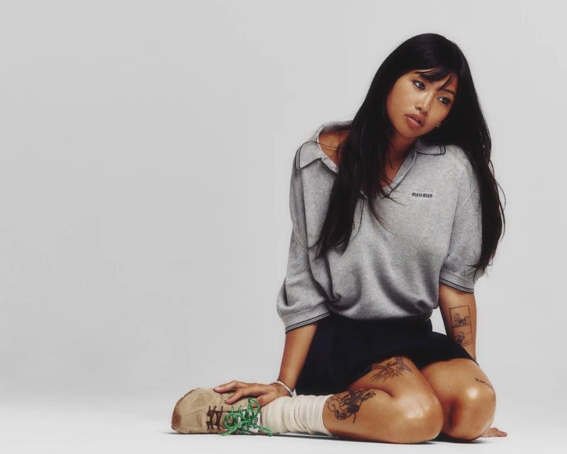 Asian woman in gray sweatshirt and navy skirt, kneeling indoors with tattoos, tan sneakers, and cream socks against a gray background.