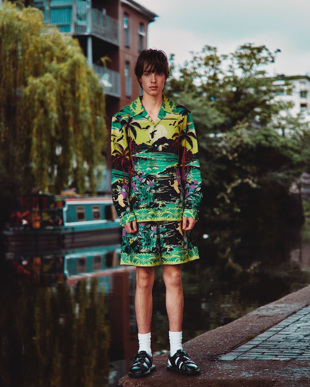 Teenage male fashion model in tropical print matching short-sleeve shirt and shorts standing on brick sidewalk by canal.