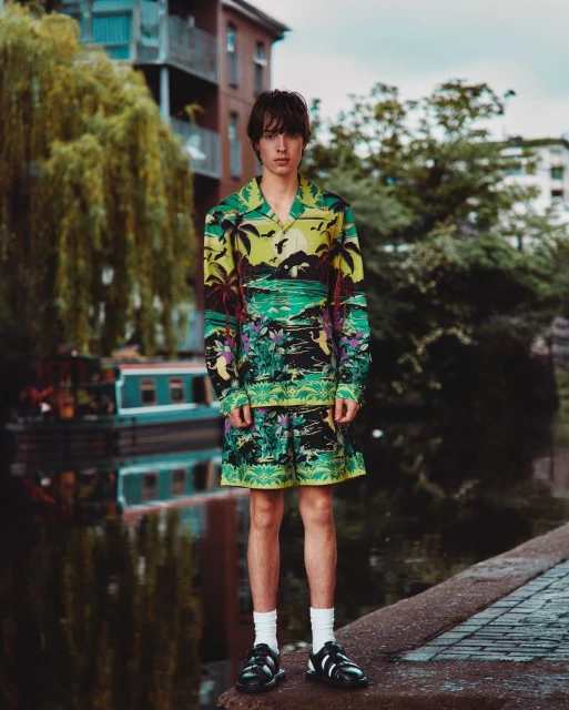 Teenage male fashion model in tropical print matching short-sleeve shirt and shorts standing on brick sidewalk by canal.