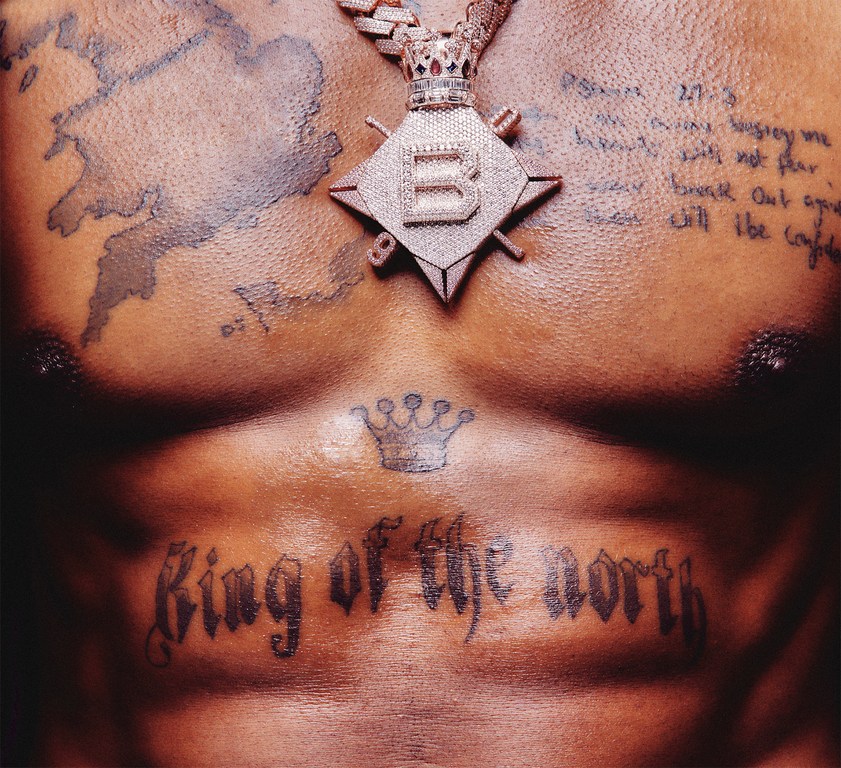 Close-up shot of a dark-skinned man's chest adorned with tattoos and a large diamond pendant, emphasizing texture and luxury.