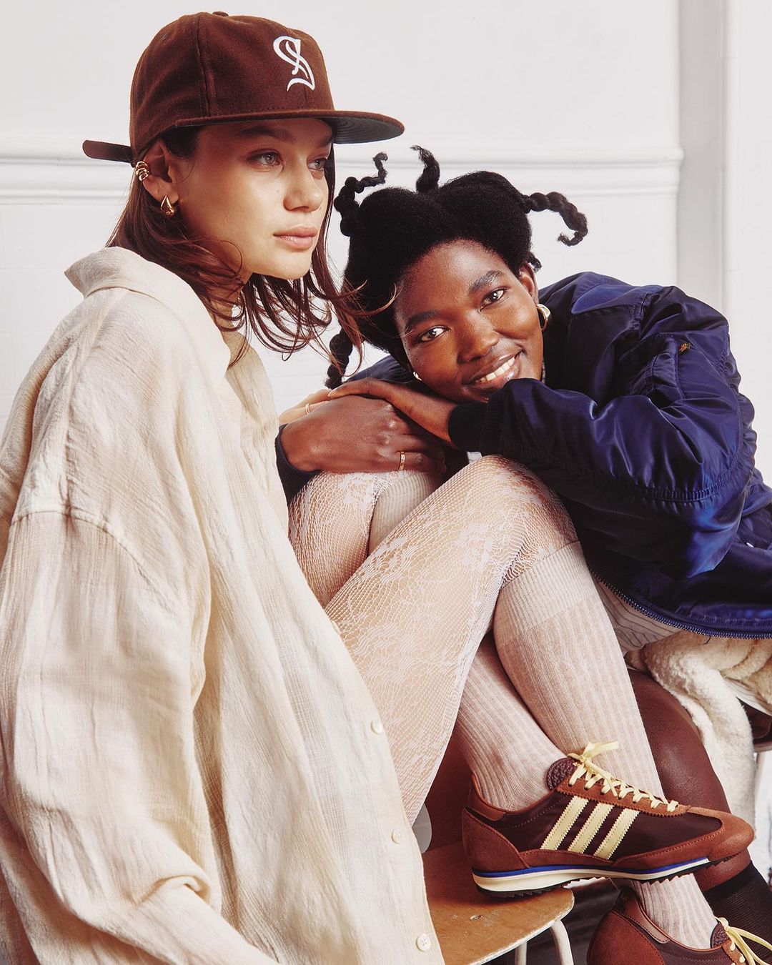 Two diverse young adult women pose together indoors. One wears a brown cap and cream shirt; the other wears a blue bomber jacket and brown sneakers.