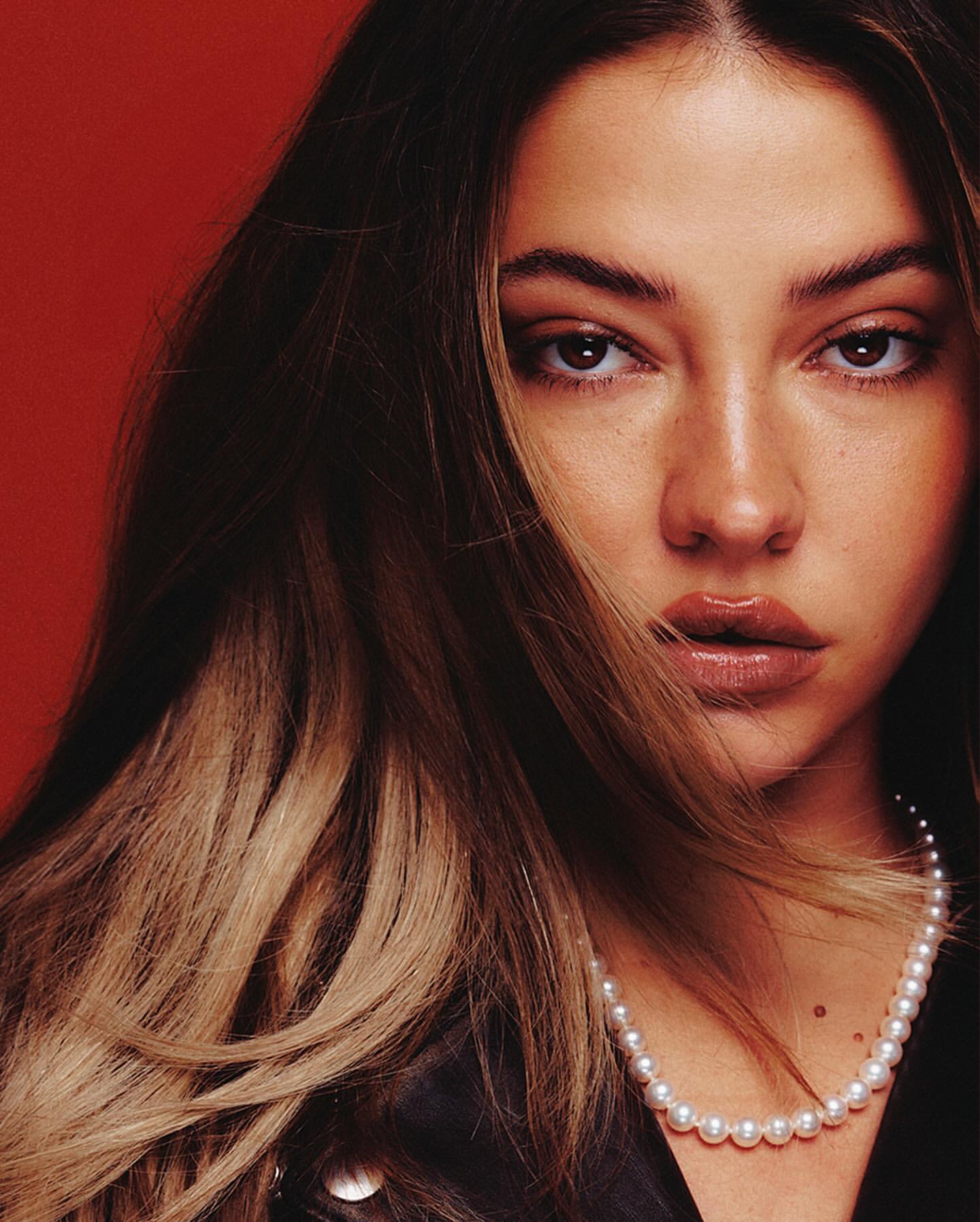 Close-up portrait of a woman with ombre hair and a pearl necklace against a vibrant red background in a studio setting.