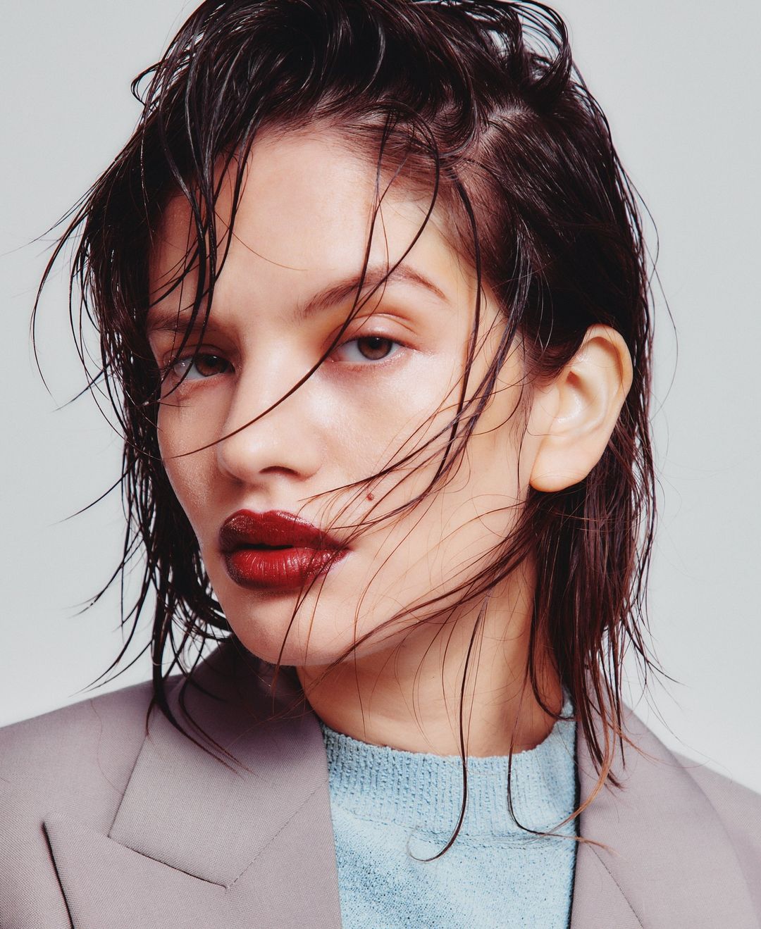 Close-up portrait of a woman with wet dark hair and red lipstick, wearing a light blue sweater and grey jacket against a neutral background.