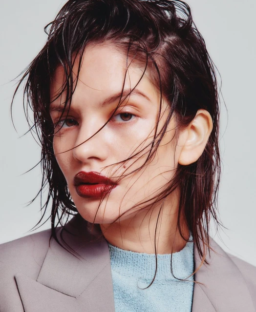 Close-up portrait of a woman with wet dark hair and red lipstick, wearing a light blue sweater and grey jacket against a neutral background.