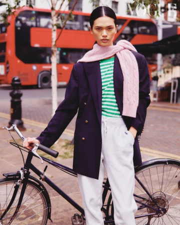 An adult Asian female poses with a bicycle on a city street, wearing a blazer, striped shirt, sweatpants, and scarf, Vogue magazine.