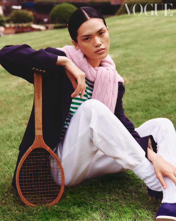 A person sits on grass with a tennis racket, wearing a striped shirt, scarf, navy jacket, and white pants, styled for a fashion editorial.