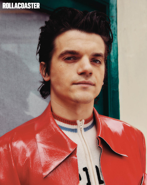 Indoor portrait of a fair-skinned young adult male with dark hair wearing a red patent leather jacket and patterned knit sweater.