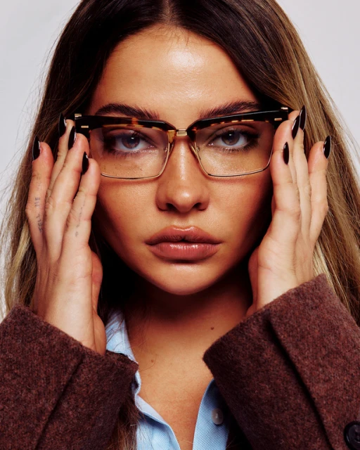 Close-up portrait of a fair-skinned adult female model with long brown hair and glasses, wearing a brown jacket and blue blouse.