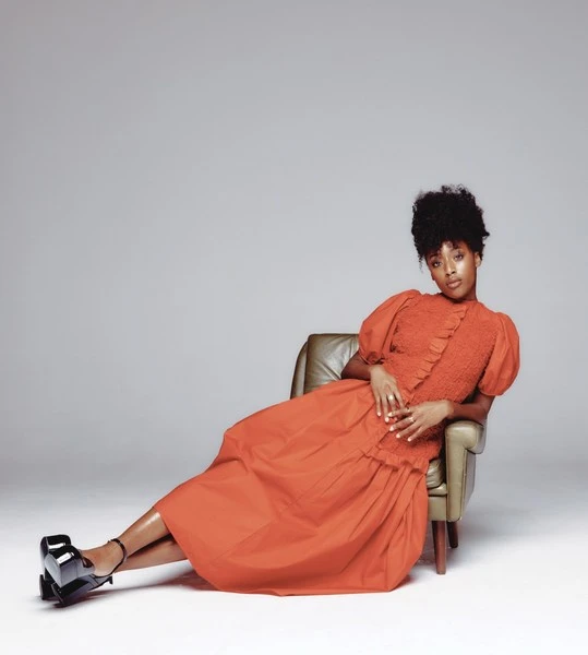 Portrait of a Black woman in a terracotta dress, reclining in a chair. The model wears black platform shoes and poses in a photo studio.