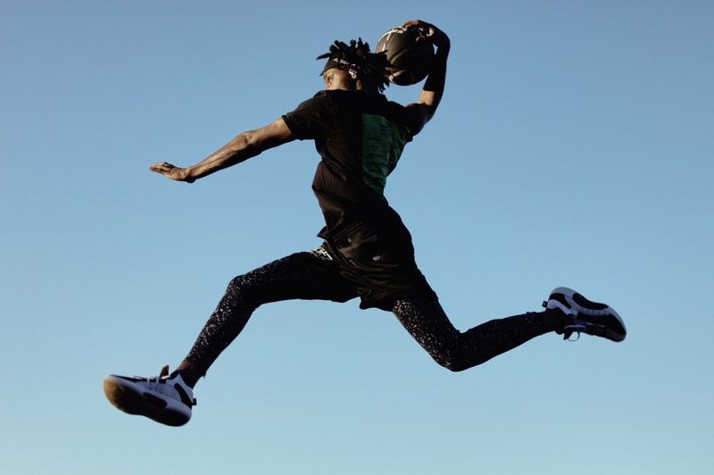 Man in mid-air about to dunk basketball against a blue sky. The athlete wears a black t-shirt, shorts, patterned leggings and athletic shoes.