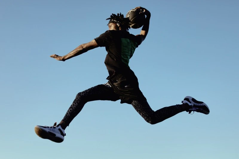 Man in mid-air about to dunk basketball against a blue sky. The athlete wears a black t-shirt, shorts, patterned leggings and athletic shoes.