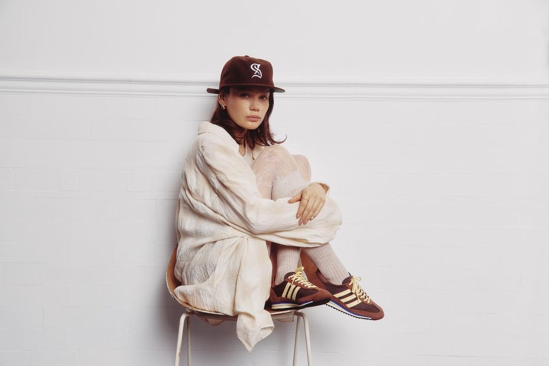A young woman with a cap and sporty streetwear sits on a metal stool in front of a white wall; brown and yellow Adidas sneakers, white socks.