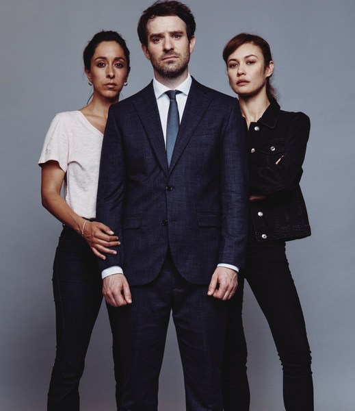 Studio shot featuring a man in a suit flanked by two women in dark clothing against a gray background. The man is centered.