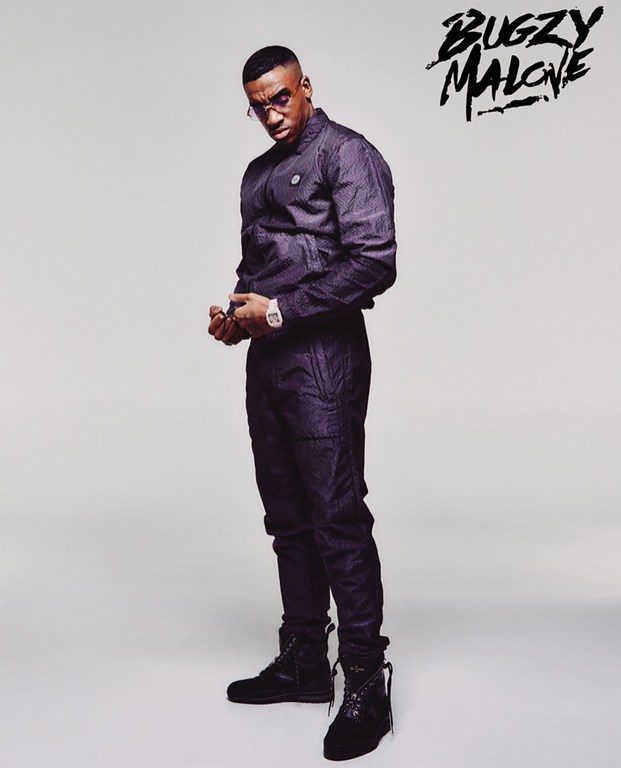 Portrait of British rapper Bugzy Malone wearing a dark tracksuit and boots against a white studio background.