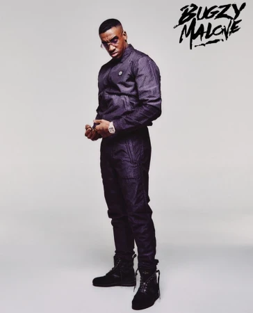 Portrait of British rapper Bugzy Malone wearing a dark tracksuit and boots against a white studio background.