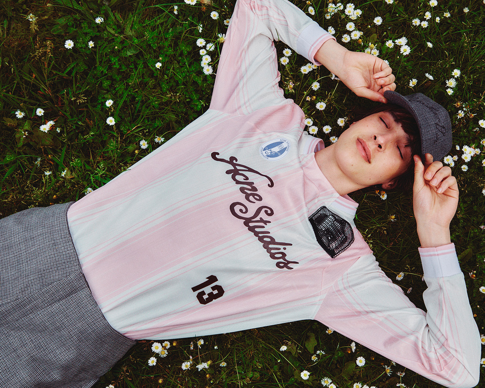 Young adult male laying in grass with daisies wearing a pink and white long sleeve Acne Studios shirt and a gray cap outdoors.