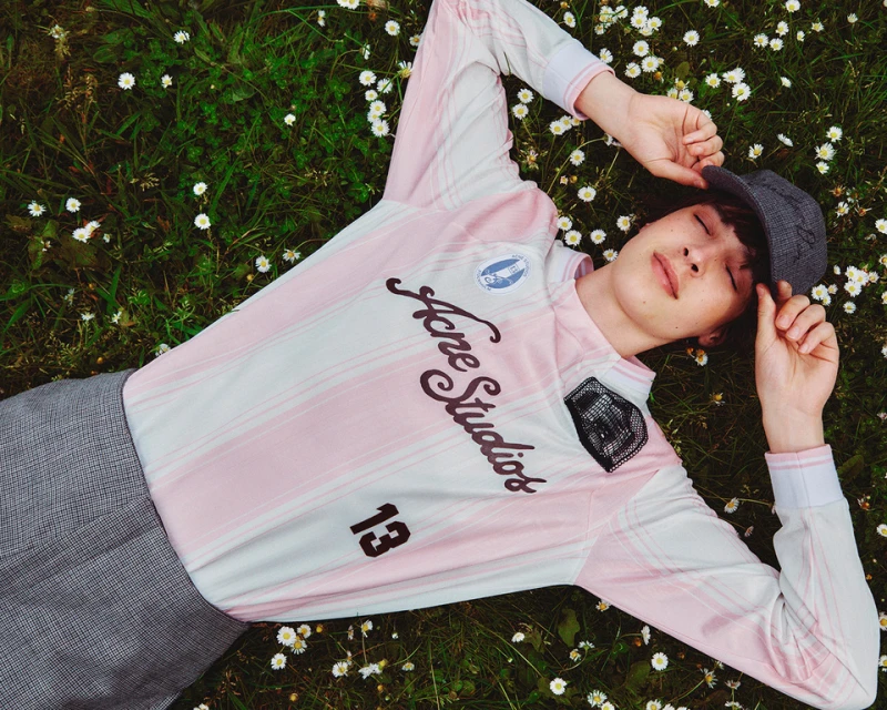 Young adult male laying in grass with daisies wearing a pink and white long sleeve Acne Studios shirt and a gray cap outdoors.