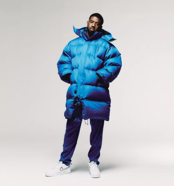 Full-length shot of adult male wearing a blue puffer coat and matching pants, accessorized with white sneakers, set against a white backdrop.