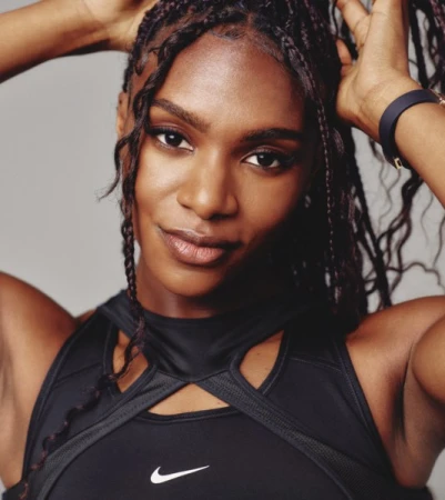 Portrait of a Black female athlete with hands in her hair, wearing a black sports bra with a white logo, against a grey background.