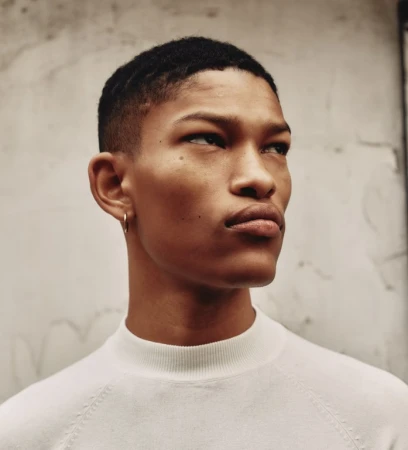 Close-up portrait of a young man wearing a white textured sweater and small hoop earring against a neutral wall