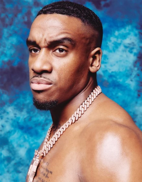Close-up studio portrait of a serious adult male with a shaved head and a gold chain against a mottled blue and white background.