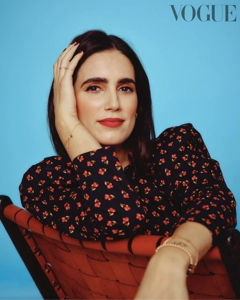 Portrait of a brunette woman in a patterned dress seated on a red chair against a light blue background, gazing at the camera.