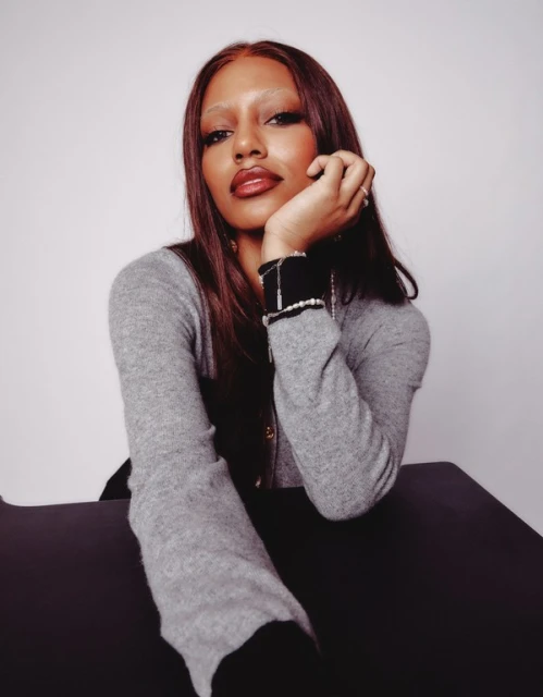 Portrait of an adult woman with long brown hair wearing a gray cardigan, resting her chin on her hand, posing in a photo studio.