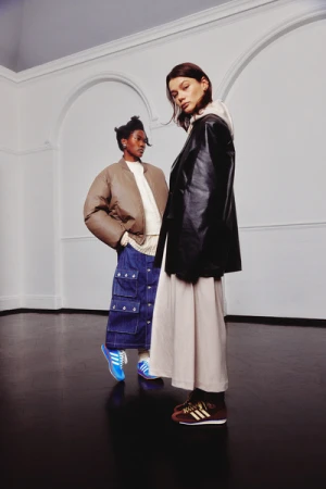 Two stylish people pose in front of a white wall wearing a brown jacket, a black leather jacket, a white top, blue jeans, and sneakers.