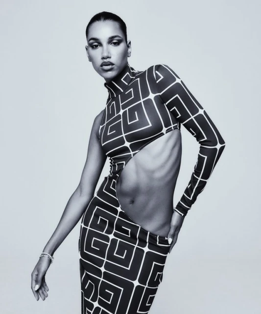 A woman in a geometric-patterned one-shoulder dress poses in a studio with a neutral background, showcasing fashion in a black and white photograph.