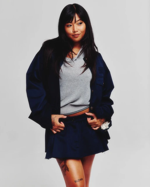 Young woman with long dark hair poses in a gray top, blue jacket, and blue skirt against a plain white backdrop in a studio shot.