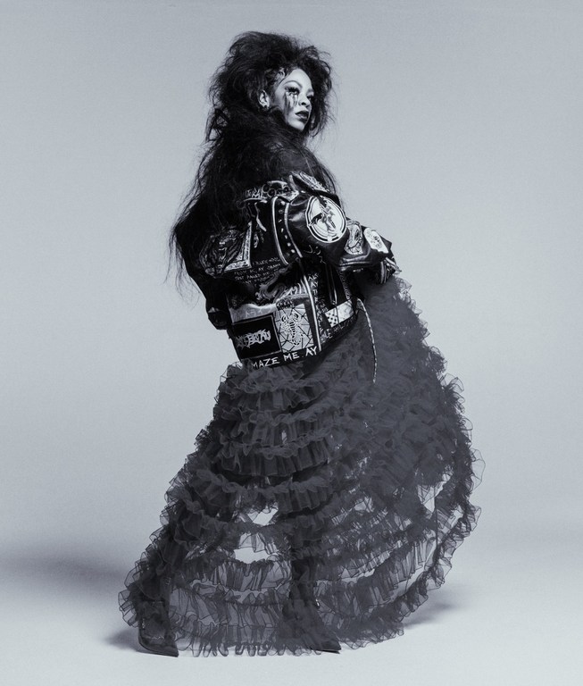 A person with dark hair and makeup in a black and white image wearing a jacket and black ruffled skirt posing in a studio setting.