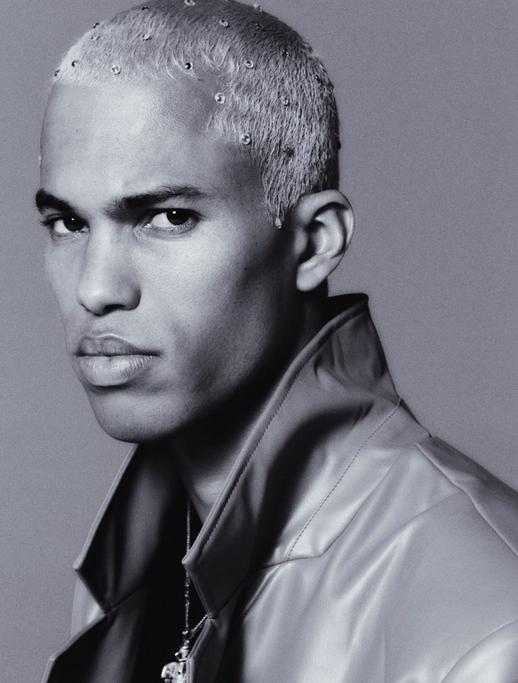 Close-up studio portrait of a light-skinned adult male with buzzed, light blonde hair and multiple metal piercings wearing a metallic jacket.