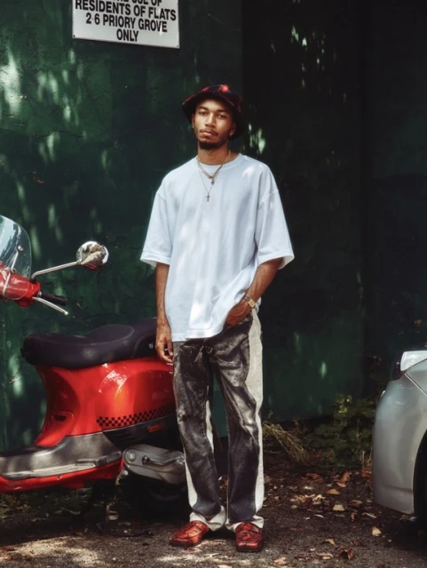 Young man posing next to a red motor scooter and parked car. He wears a white t-shirt, patterned bucket hat, and dark wash jeans.