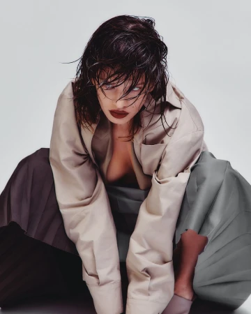 A woman with wet dark hair wearing a beige button-down shirt and grey skirt against a white background by Ryan Saradjola