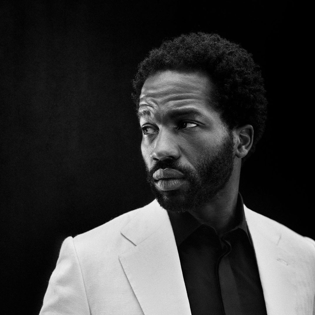 Monochrome portrait of an African American man in a white suit jacket looking off to the left against a dark background.