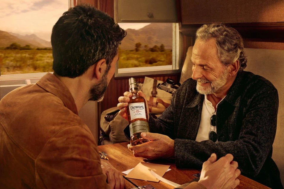 Two men inside a train car enjoy a bottle of Dewar's 15-year-old scotch whisky, showcasing a lifestyle moment with warm lighting and a scenic view.