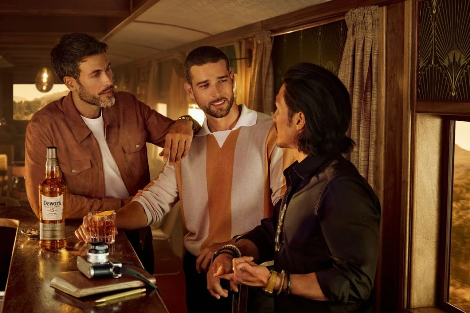 Three men in a relaxed, indoor setting, featuring a bottle of Dewar's scotch, a cocktail glass, and a vintage camera on a bar. 