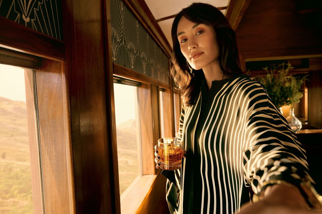 Woman in striped top holds drink in vintage train with scenic landscape view, capturing a blend of elegance and travel ambiance.